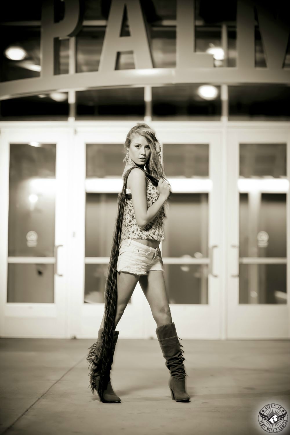 blue eyed  curly blond haired female model wearing a flowery light colored tank top and white cut off jeans in boots with tassels in front of the palmer events center entrance in a austin fashion photography location